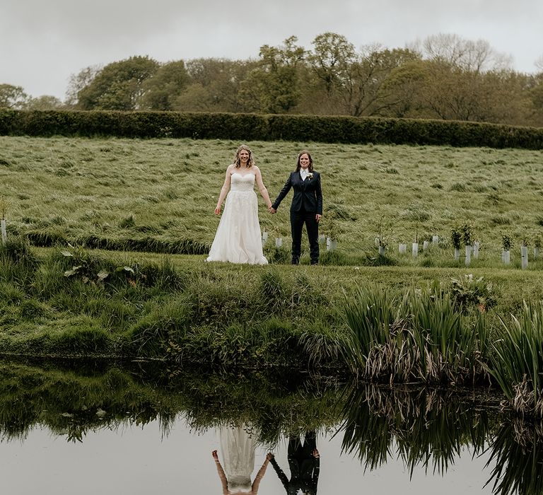 bride in a  strapless lace wedding dress and bride in a navy bridal suit holding hands in front of a lake at Stanford Farm wedding venue 