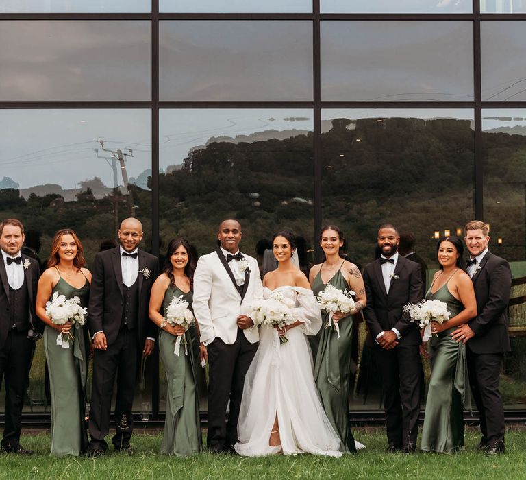 Wedding party in black tuxedos and green satin bridesmaid dresses stand in front of large window at crumplebury wedding