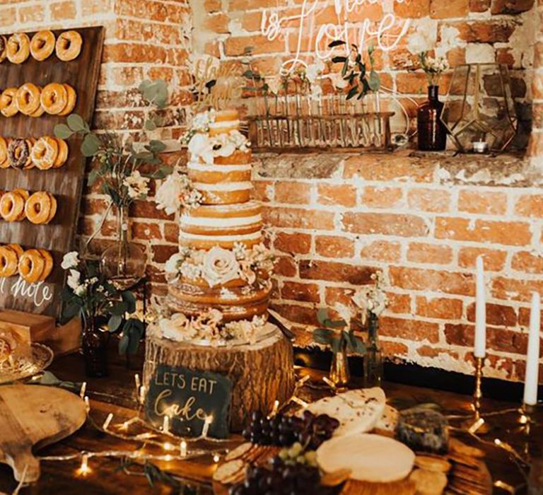 Naked wedding cake at rustic wedding on tree trunk cake stand with dessert table full of doughnuts, cheeses and more 