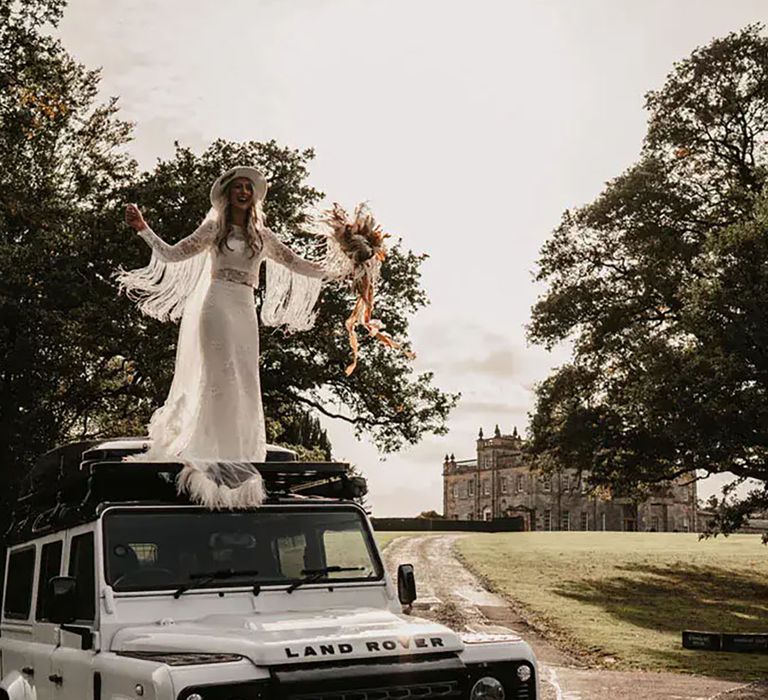 Boho bride standing on Land Rover wearing cowboy boots and a white bridal cowboy hat by Willow & Wilde Photography