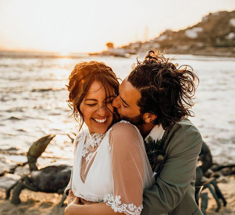 Bride wearing wedding dress with embroidered lace cape being embraced by the groom in a green suit on the beach 