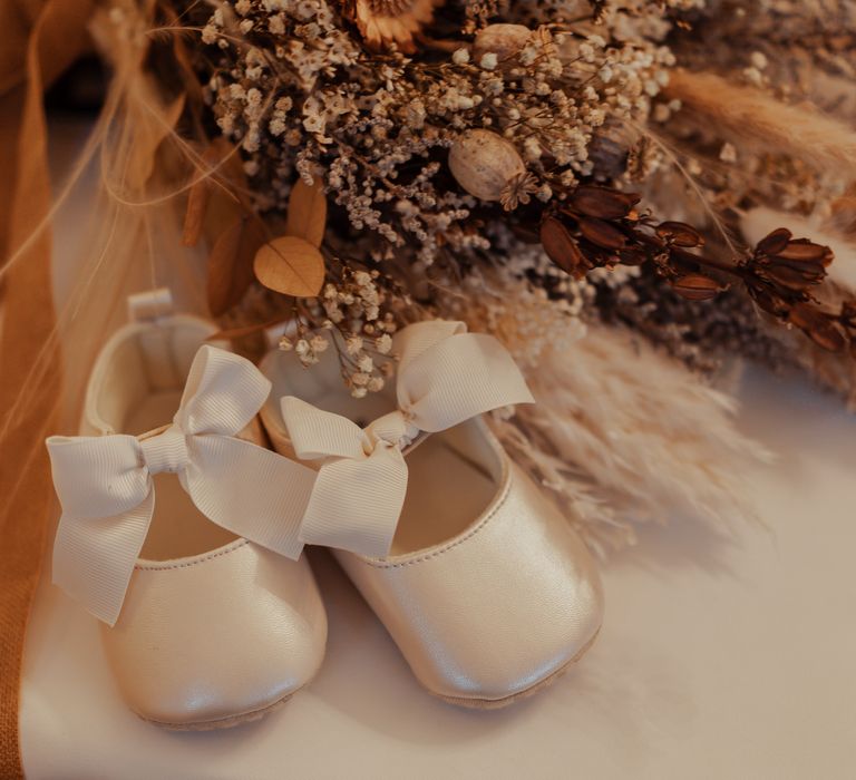 Silk flower girl shoes with ribbon tie next to dried floral bouquet 