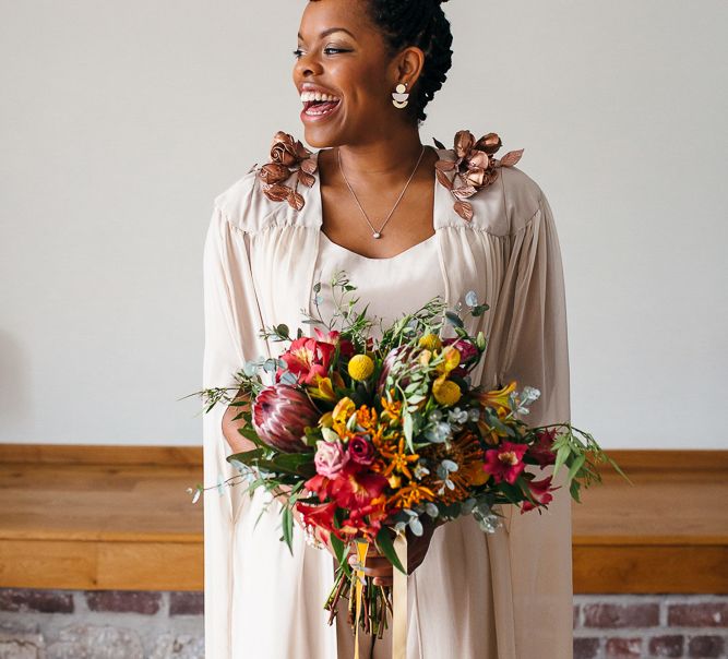 A bride wears long braids in a high bun as an example of wedding hairstyles for Black brides. Her dress has a cape and she hold a bouquet.