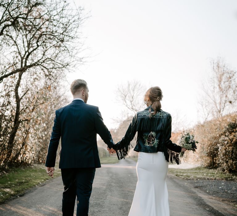 Bride in a fitted wedding dress, bridal up do and customised leather jacket with fringe detail walking up a country land with her husband 