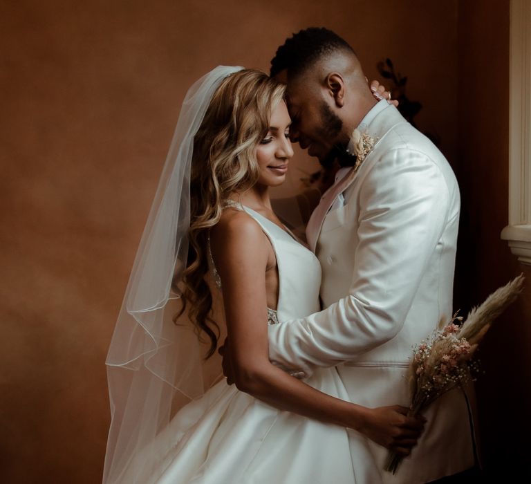 Groom in a white tuxedo embracing his bride with side swept wavy hair holding a dried flower bouquet 