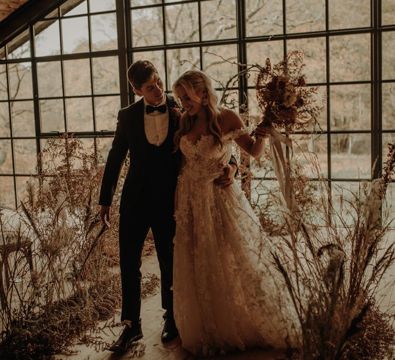 Groom in a horseshoe waistcoat with bow tie walking up the dried flower aisle with his bride in a lace wedding dress at Hidden River Cabins wedding venue 