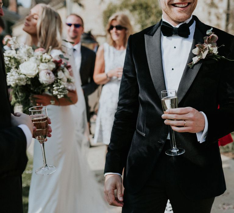 Smiling groom in black velvet Hugo Boss suit with black bow tie holding glass of sparkling white wine