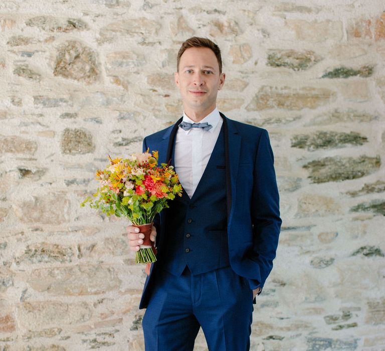 The groom holding a bouquet, wearing a dark blue three piece suit