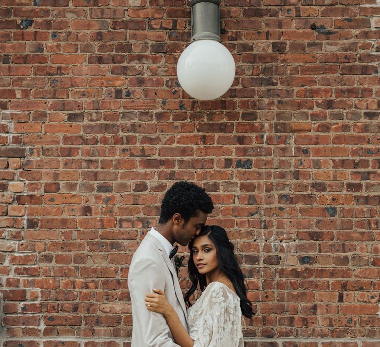 Groom in a grey blazer kissing his bride on the head in a long sleeve mermaid wedding dress with embellished detail