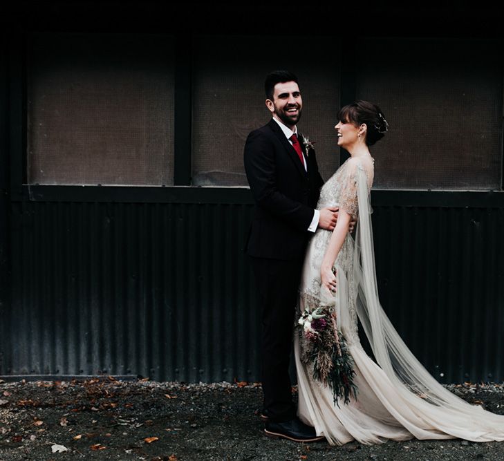 A bride and groom laugh with each other, She holds an oversized homemade wedding bouquet.