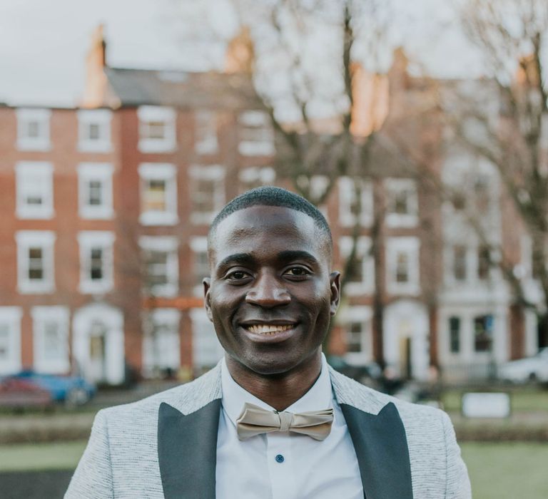 Smiling groom in grey Moss Bros suit jacket with white shirt, black waistcoat and gold satin bow tie 