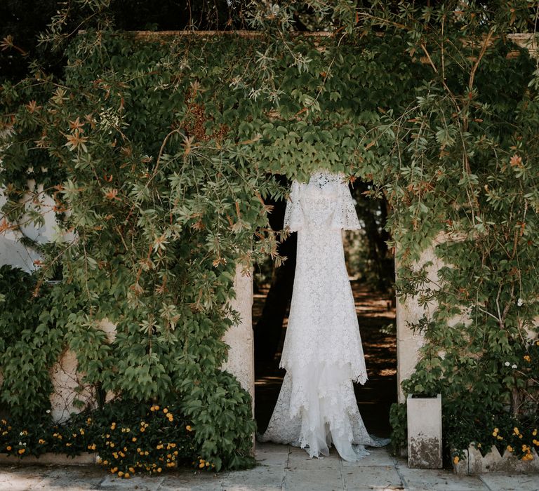 Lace boho wedding dress hangs in doorway outside between greenery at enchanted forest wedding in Italy