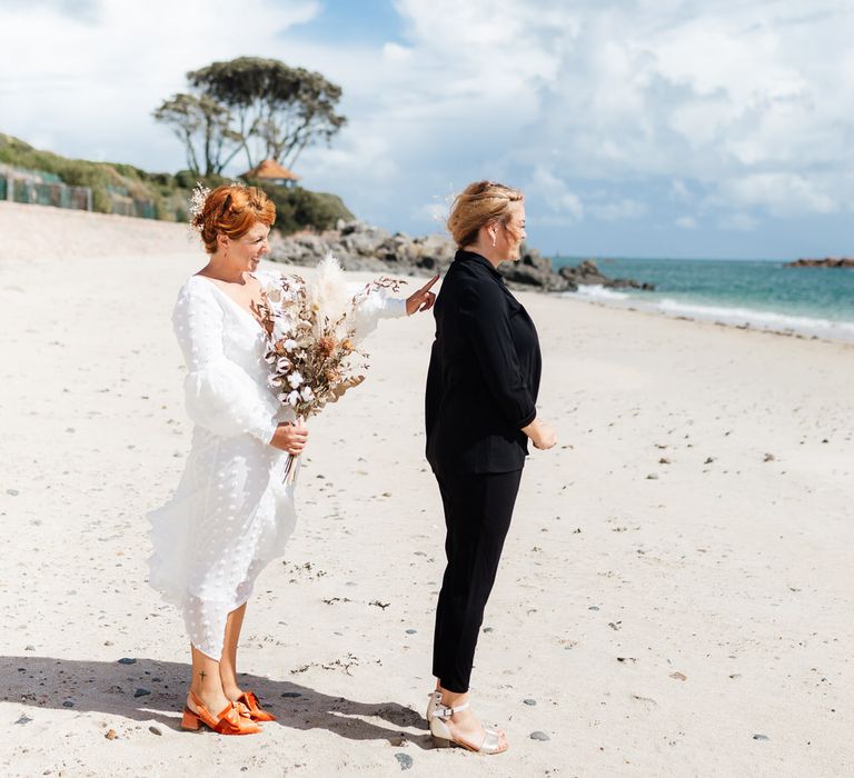 Bride in dress standing behind bride in suit on beach for first look shot