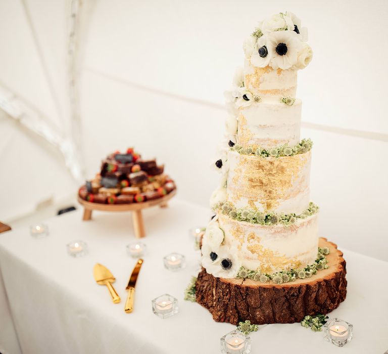 Four tiered wedding cake with white and gold detailing atop a tree slice cake stand with white and green floral decor