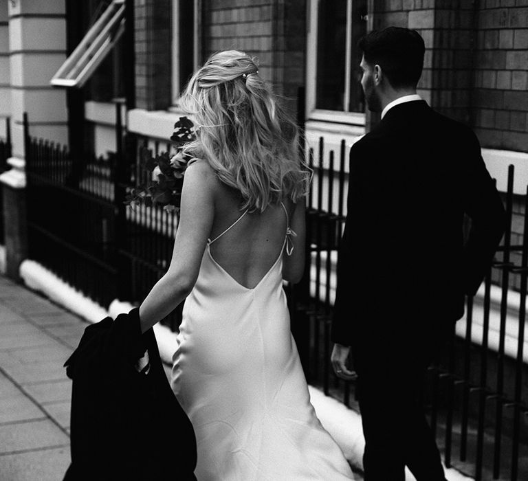 Bride with half up half down hair walking through the streets in a backless satin wedding dress with her skirt blowing in the wind 