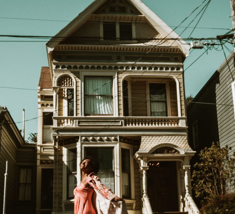 Bride-to-be in a coral dress and long-line patterned coat dancing in front of a house in San Francisco 