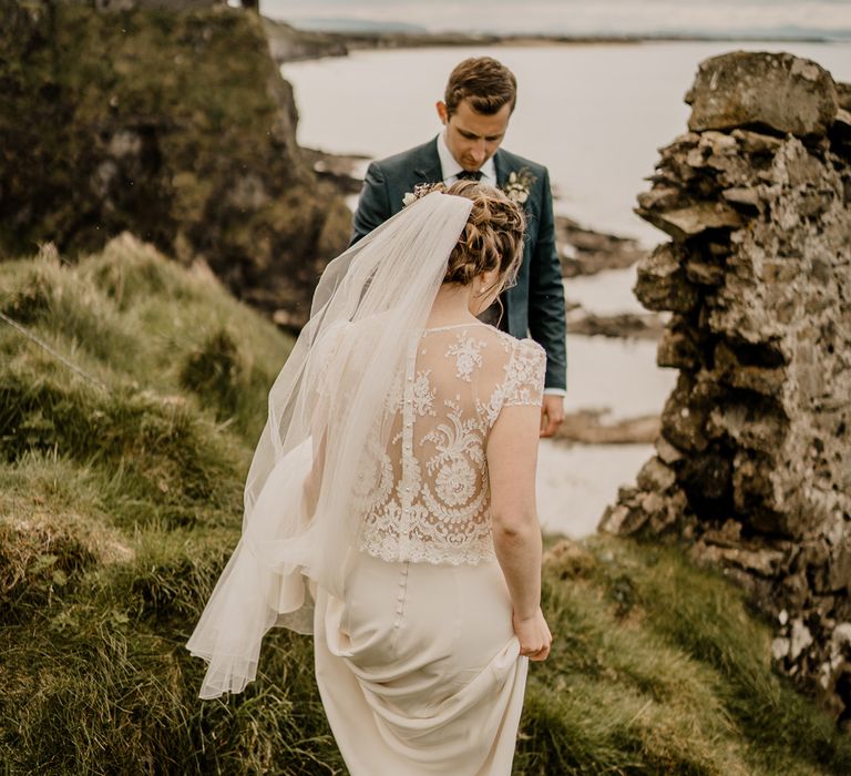 Bride in lace top wedding dress with capped sleeves and satin skirt and veil walks along cliffside towards groom in navy suit at Dunluce Castle wedding 