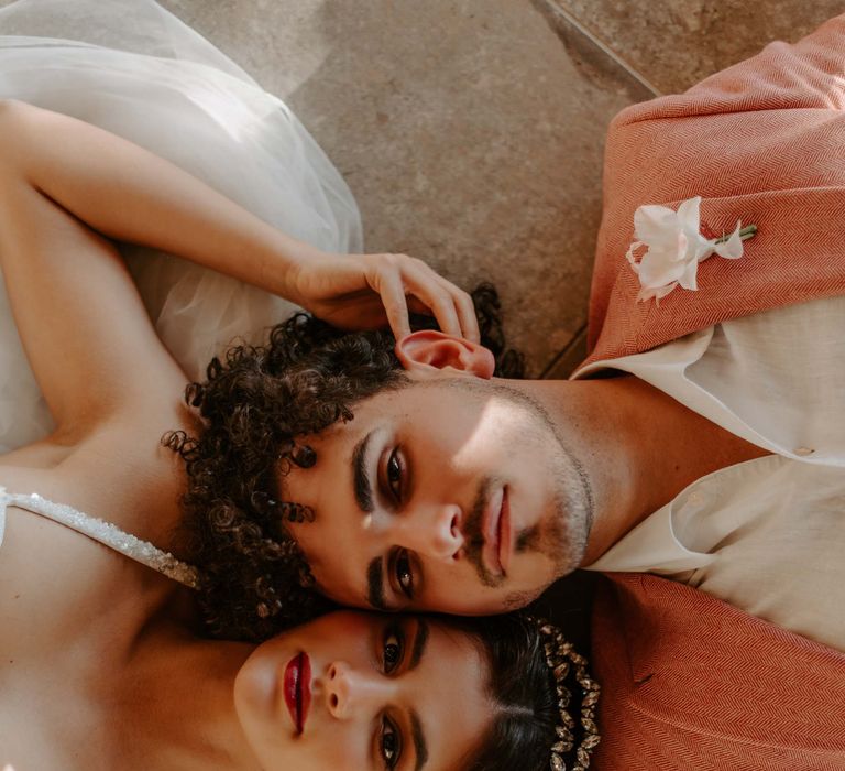 Groom with curly hair in a pink suit jacket laying on the floor with his bride 