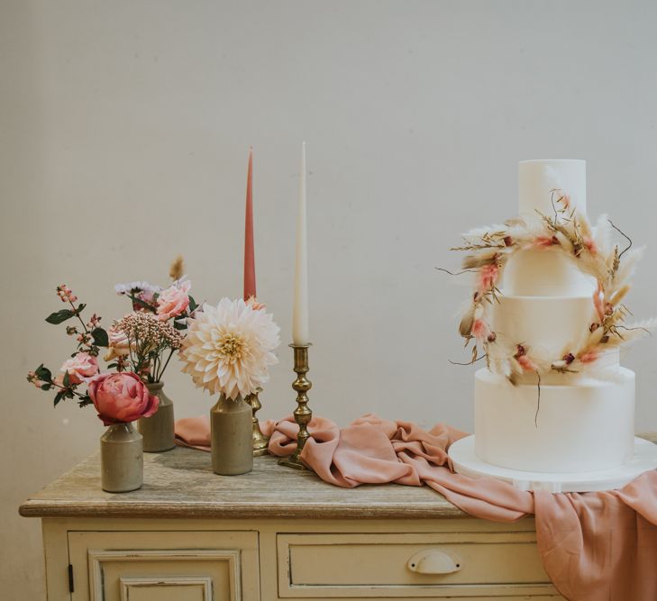 White iced wedding cake with pink and dried grass floral hoop decor on a vintage dresser surrounded by pink linens, and flower stems in vessels