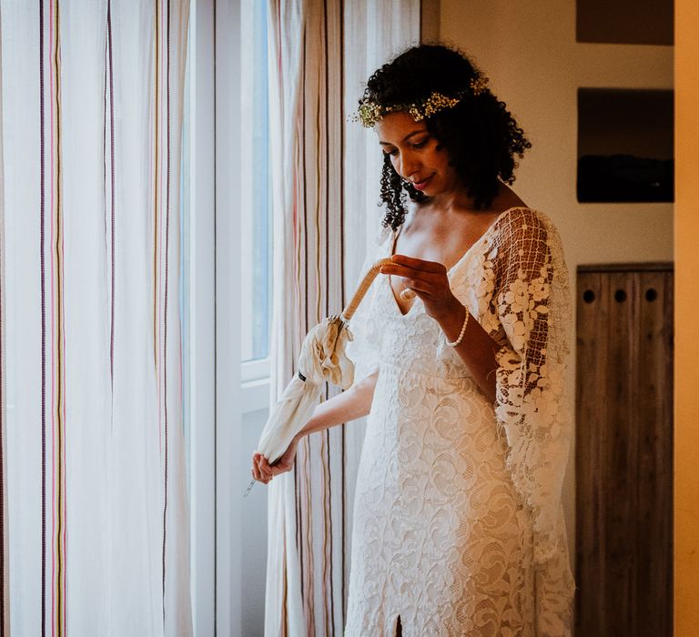 Bride looks at umbrella for her wedding day whilst wearing Grace Loves Lace wedding gown and stand beside window