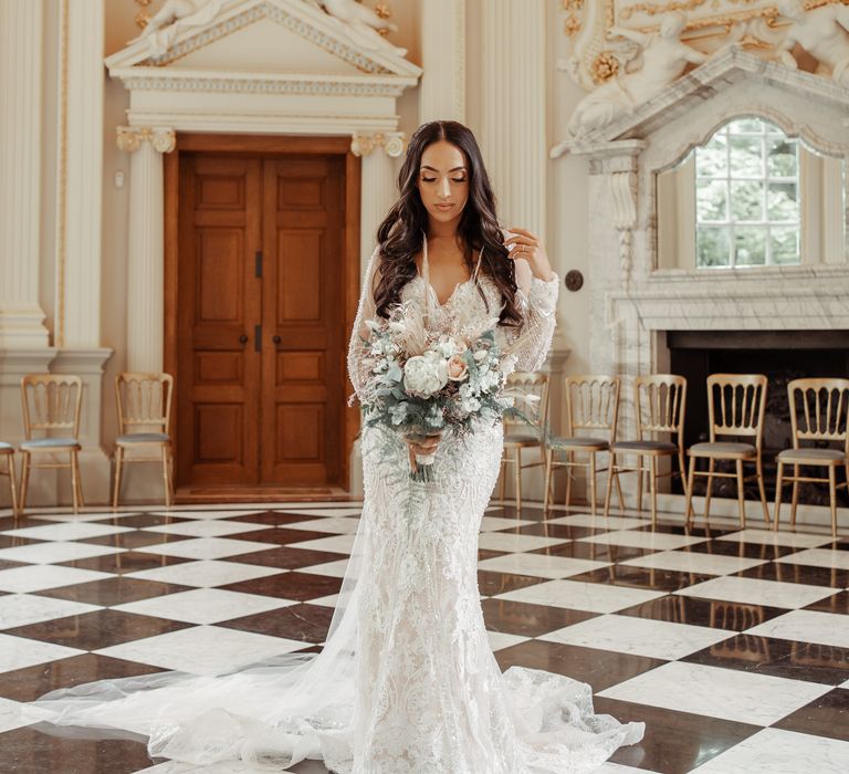 Bride stands within regal room wearing Milla Nova wedding gown and wears her long dark hair in waves