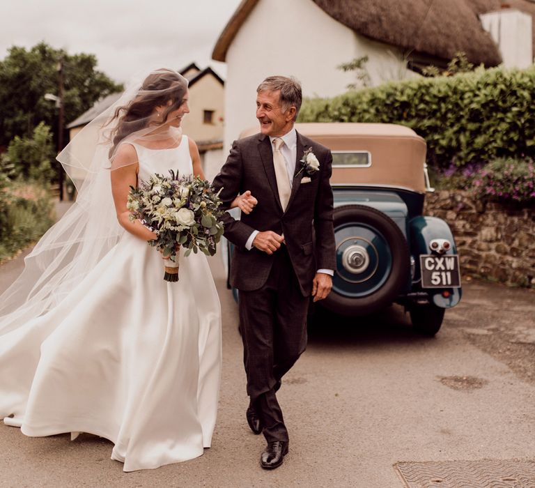 Bride in satin Elbeth Gillis wedding gown, long veil and white and green bridal bouquet walks arm in arm with father in brown suit before wedding