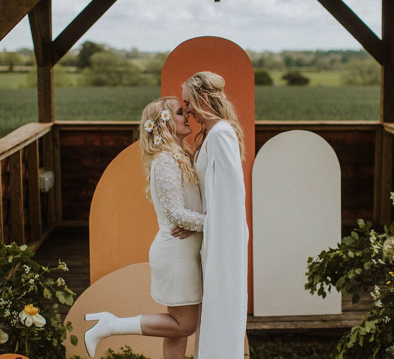 Bride in a mini wedding dress with applique long sleeves and white boots kissing her bride in a wedding jumpsuit at an outdoor sixties wedding dress