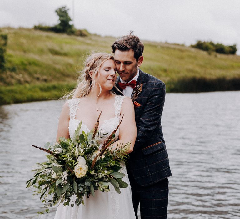 Groom wraps his arms around bride as they stand together on the jetty in front of the lake as bride carries white floral bouquet complete with feathers