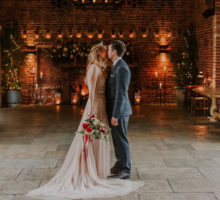 Bride in a blush tulle wedding dress with embroidered detail kissing her groom in a grey suit at their Autumn wedding with candle light 