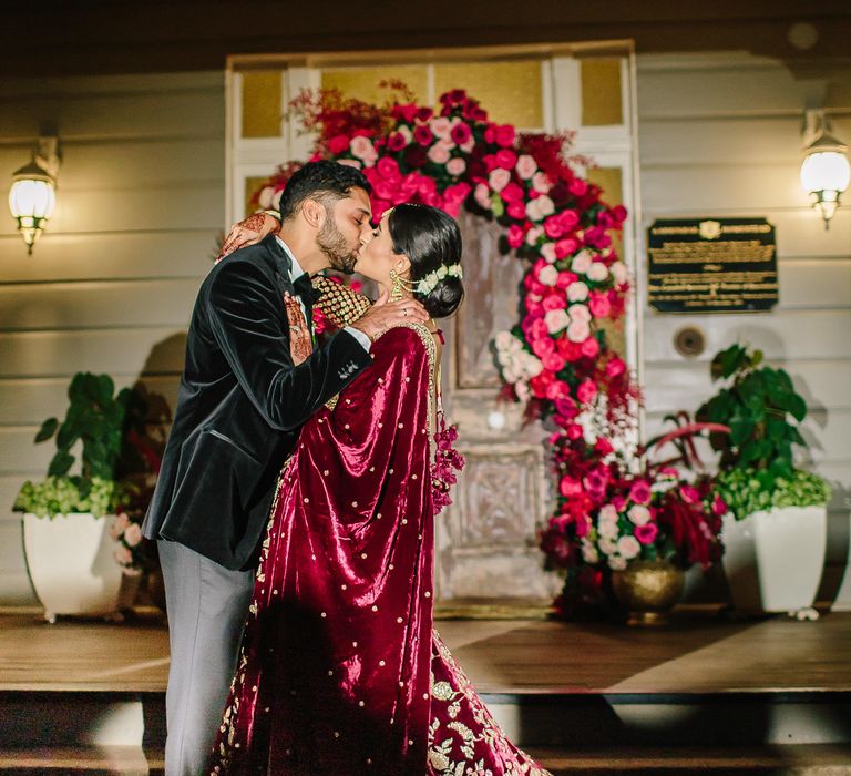 Groom kisses bride outdoors as she wears deep red lehenga on her wedding day