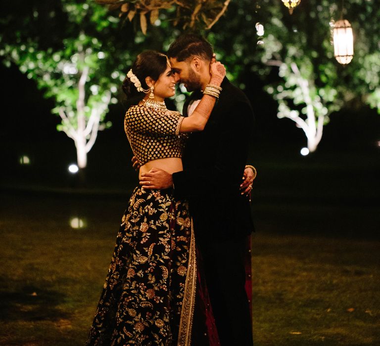 Bride & groom kiss outdoors on their wedding day as lights fill the trees and twinkle in the night