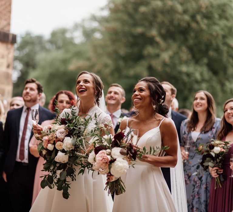 Two brides holding large white, pin and green rose bridal bouquets smile whilst looking up and holding glasses of sparkling wine whilst guests stand behind them at The West Mill Derby