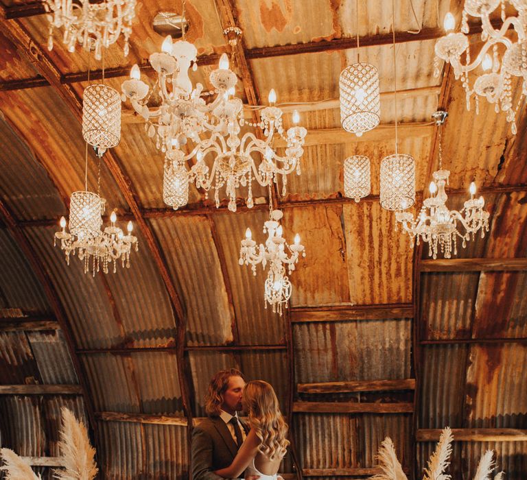 Bride in white Made With Love wedding dress with open back, button detailing and train kisses groom in sage green suit under lit chandeliers surrounded by woven baskets filled with pampas grass and dried leaves in the Hut at Anran Devon