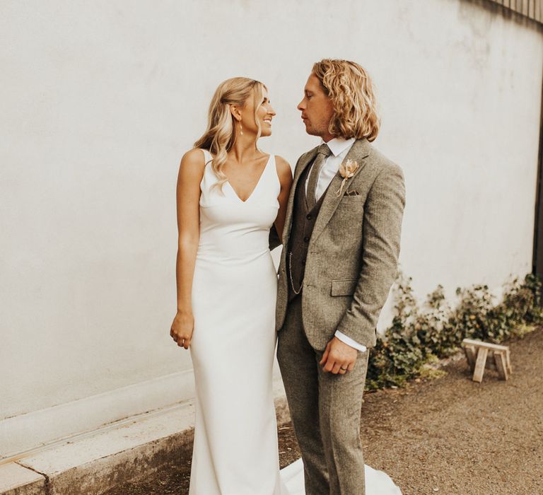 Bride in white v neck Made With Love wedding dress and groom in sage green three piece suit and brown shoes stand smiling at each other outside at Anran Devon