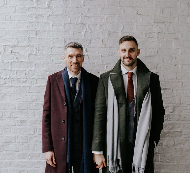 Grooms stand holding hands with one another as they look toward the camera and wear grey scarves with their suits