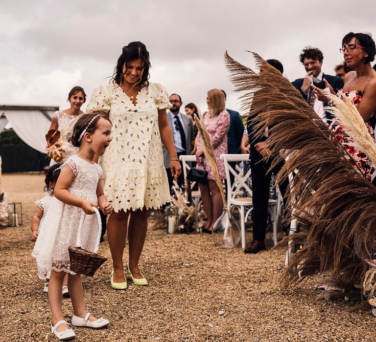 Flower girl in short lace dress holding backset walks down aisle with guests and flower girl as guests watch on during outdoor wedding ceremony in Dorset with pampas grass decor