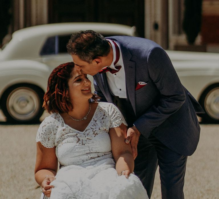 Indian groom in a three-piece blue suit with burgundy bow tie kissing his bride in a lace wedding dress on the forehead as she sits in her wheelchair 