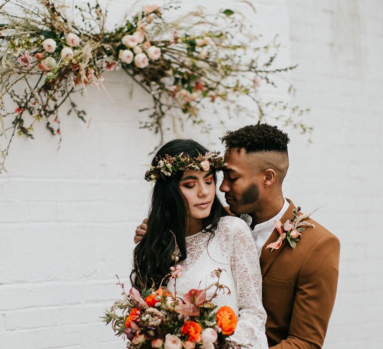 Groom in a dark orange suit embracing his bride in a lace dress and flower crown as she holds her autumnal wedding bouquet 