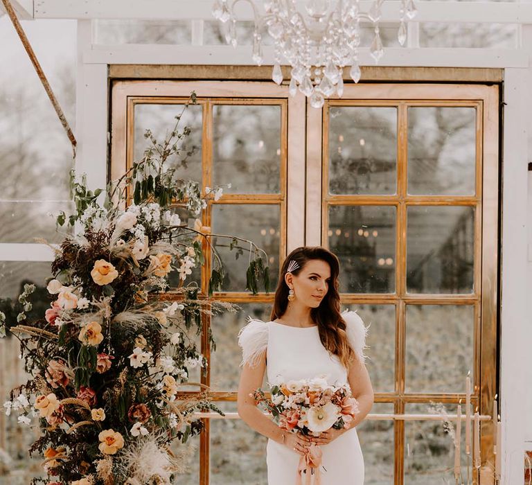 Bride with side swept hair pinned with sparkly hair grips holding her yellow and orange bridal bouquet at the altar wearing a fitted wedding dress with puddle train and feather cap sleeves