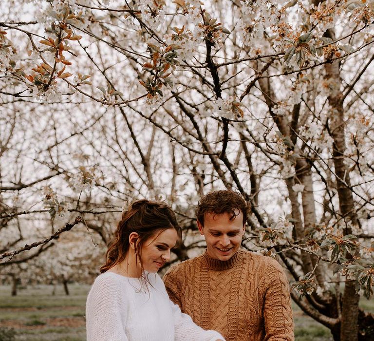 Bride in a laser cut lace wedding dress with knitted jumper cutting the wedding cake in a forest with her husband in a brown jumper 