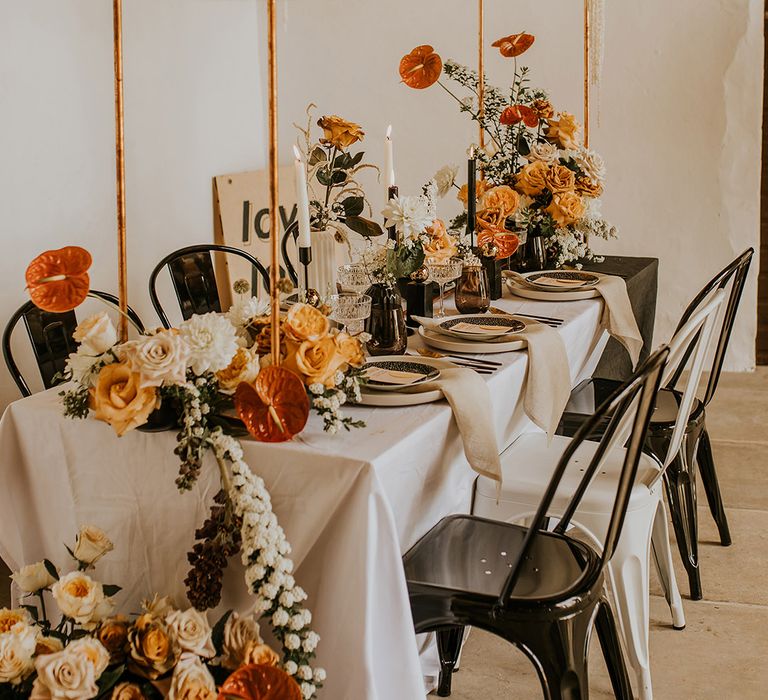 Intimate reception table decor with black industrial chairs and candles, linen tablecloth and orange and yellow flowers 