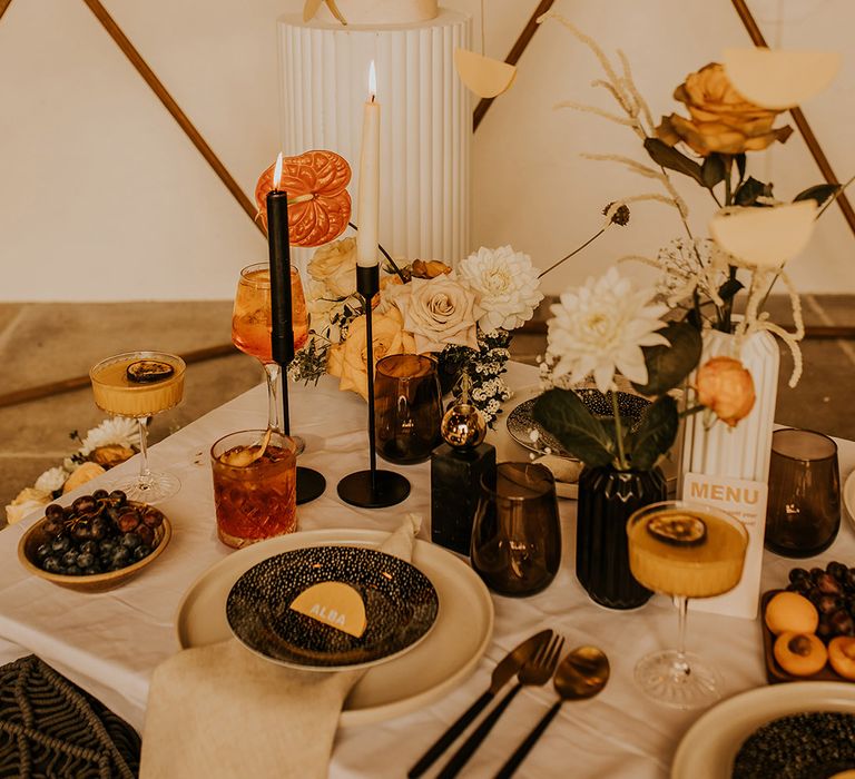 Wedding table with black tableware, cocktails, orange and yellow flowers in vases and a tall geometric marble wedding cake 