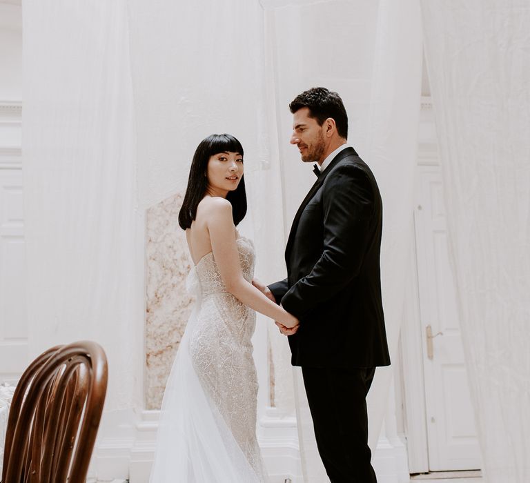 Groom in a tuxedo holding hands with his bride in a strapless embellished wedding dress with straight bob length hair and fringe 