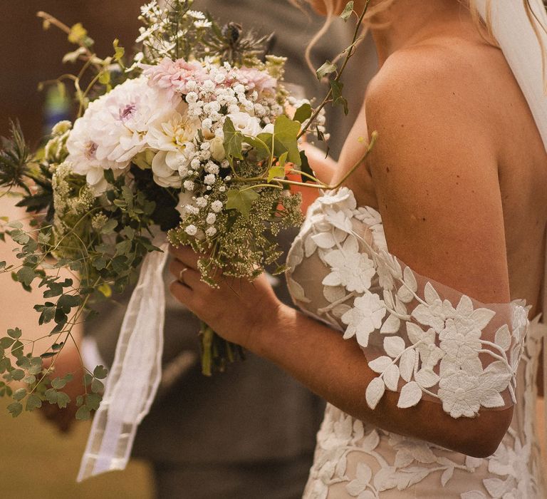 Bride in off the shoulder Enzoani wedding dress and veil holding white, pink and green wedding bouquet stands outside during tipi wedding at Inkersall Grange Farm