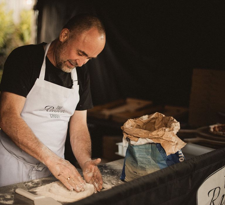 Man in black t shirt and apron makes pizza dough base by hand at tipi wedding reception during Inkersall Grange Farm wedding