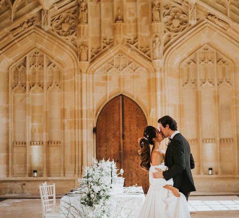 Groom in black tuxedo kissing his bride in a strapless wedding dress with full skirt and detachable sleeves at their monochrome Bodleian wedding 