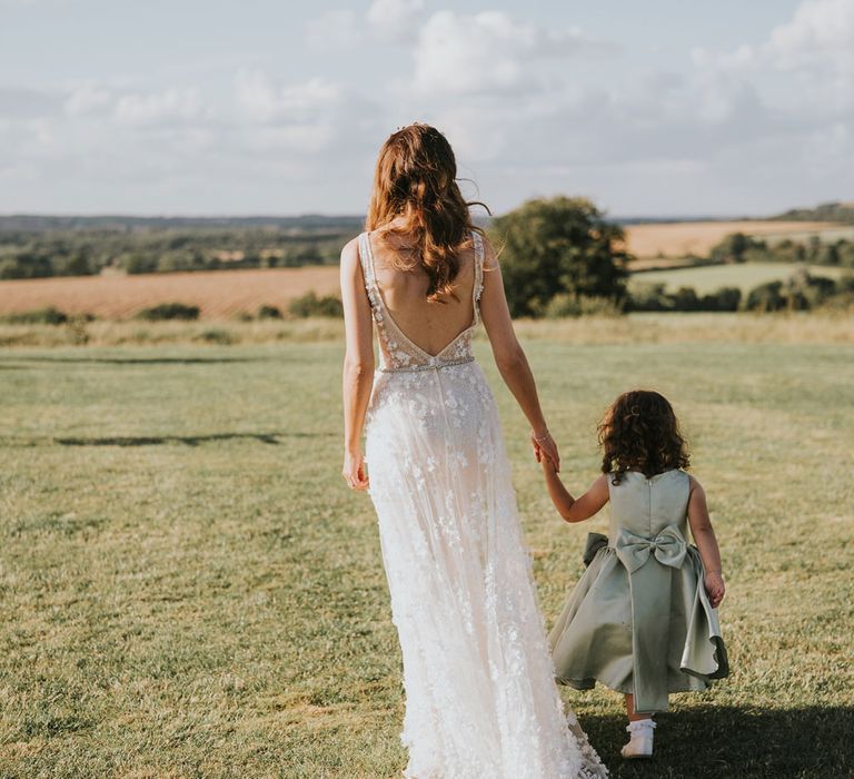 Bride in open back applique Berta wedding dress walks through field holding hands with flower girl in sage green dress with bow detail during summer wedding at Primrose Hill Farm