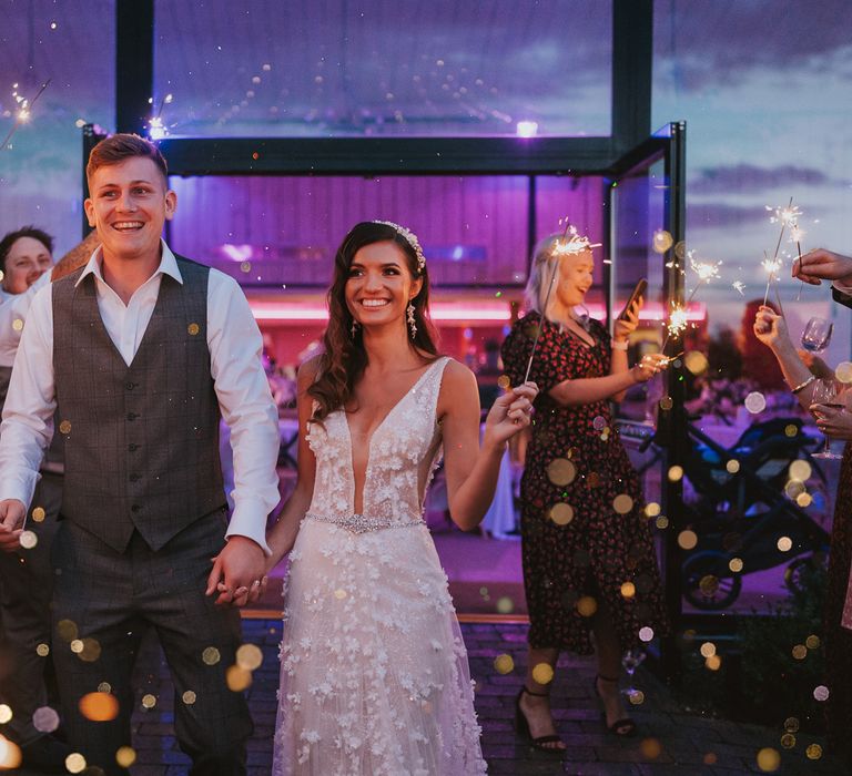 Bride in deep v applique Berta wedding dress and bridal headband walks holding hands with groom in white shirt and grey checked waistcoat for sparkler exit during summer wedding at Primrose Hill Farm