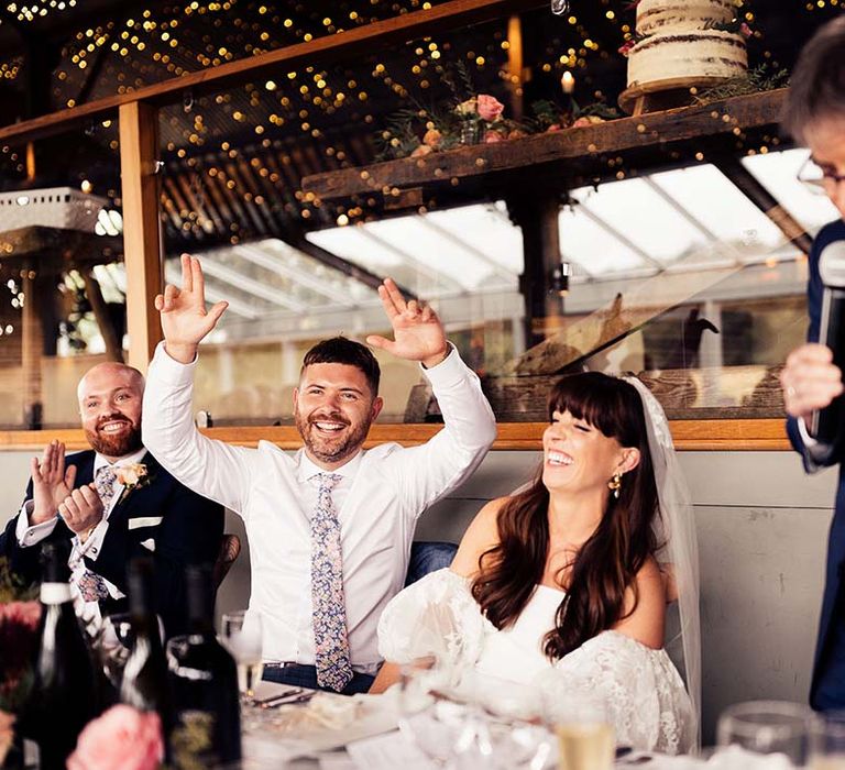 Groom in a floral tie and bride in a strapless wedding dress with lace puff sleeves laughing at the top table 