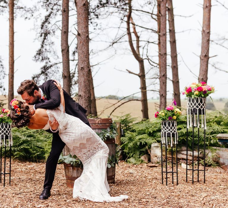 Bride in Grace Loves Lace wedding dress and flower crown is held by groom in black suit for first kiss during woodland wedding ceremony